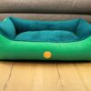 Hundebett in der Farbe türkis und grün vor einer grauen Couch auf Holzboden. Rosa Schlafplatz für Hunde.